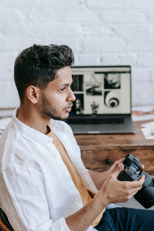 Thoughtful ethnic man with professional photo camera sitting at desk