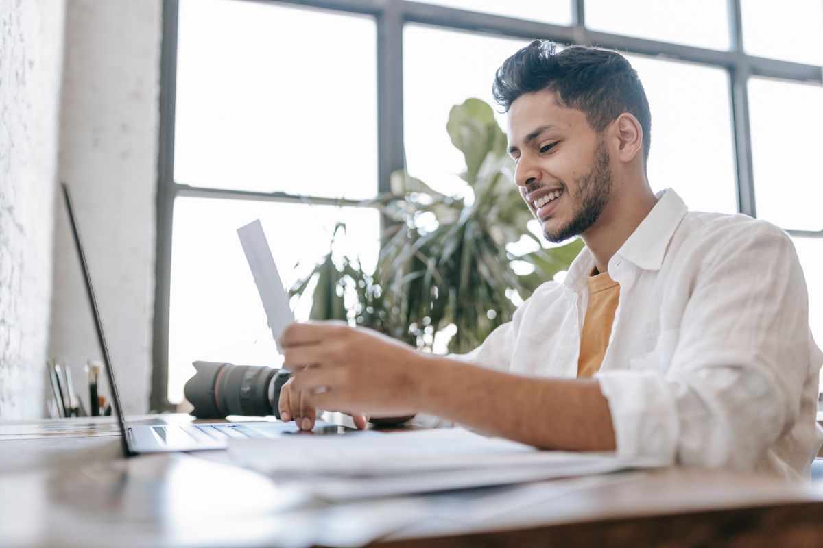 Cheerful young male looking at printed photo at desk with laptop