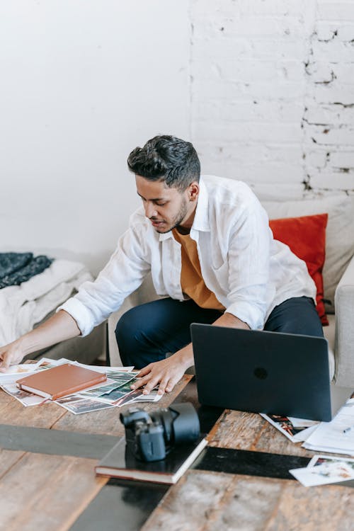 Young ethnic male photographer scattering printed documents on table near netbook and professional photo camera in living room