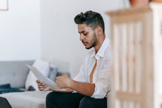 Concentrated young ethnic male wearing casual outfit reading documents and sitting on couch in modern living room