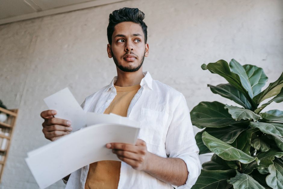 Thoughtful ethnic male in casual clothes holding printed pictures in hands and looking away while standing in light living room Thoughtful ethnic male in casual clothes holding printed pictures in hands and looking away while standing in light living room