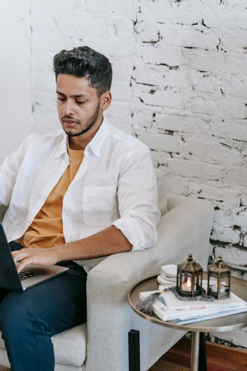 Focused ethnic man working on laptop on cozy armchair