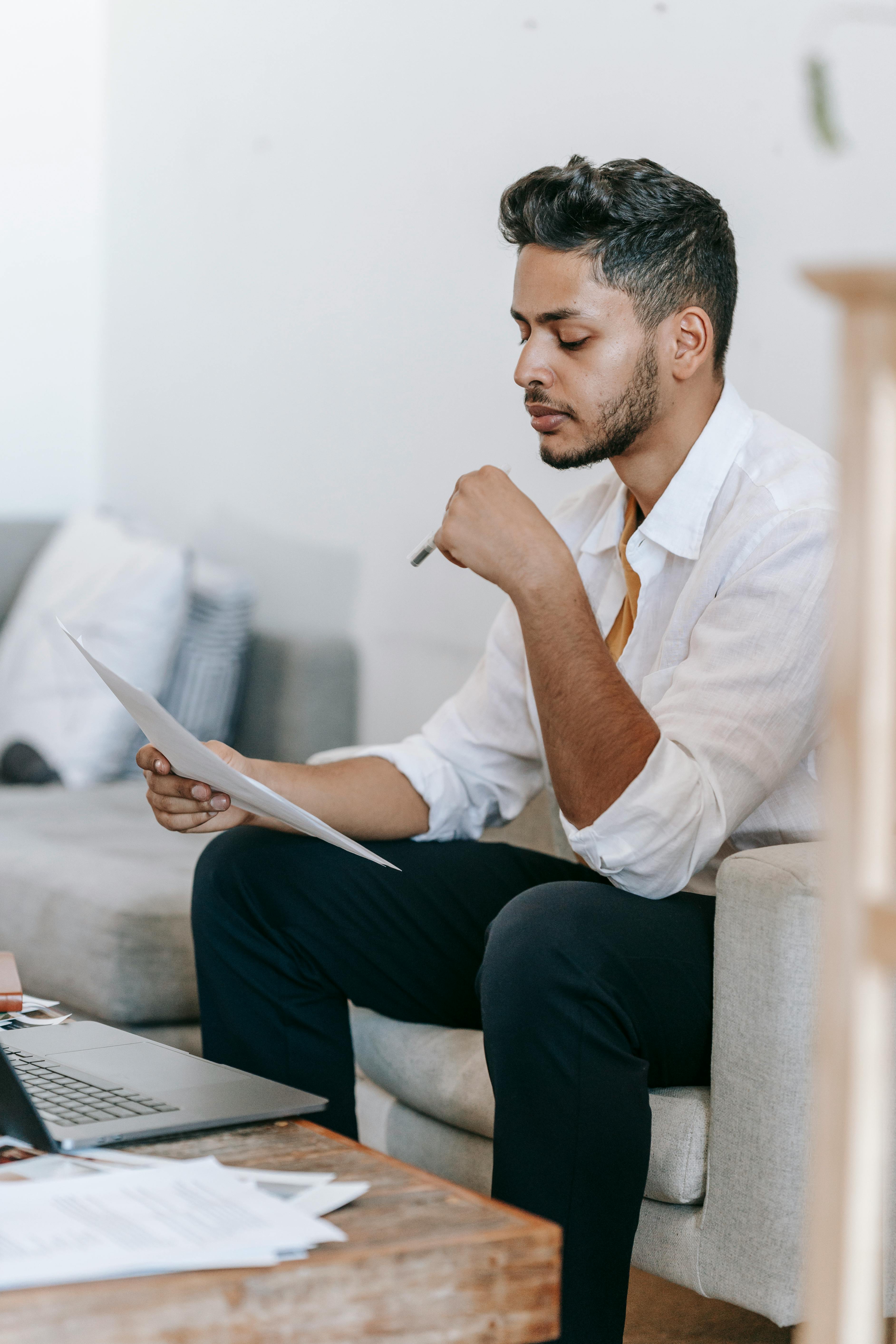 focused ethnic man reading documents and working on laptop