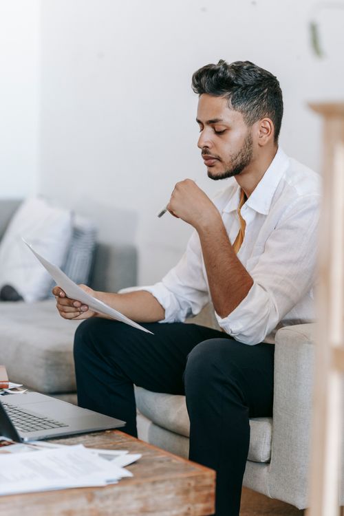 Free Concentrated young ethnic male wearing casual clothes reading documents and sitting on armchair near table with netbook in living room Stock Photo