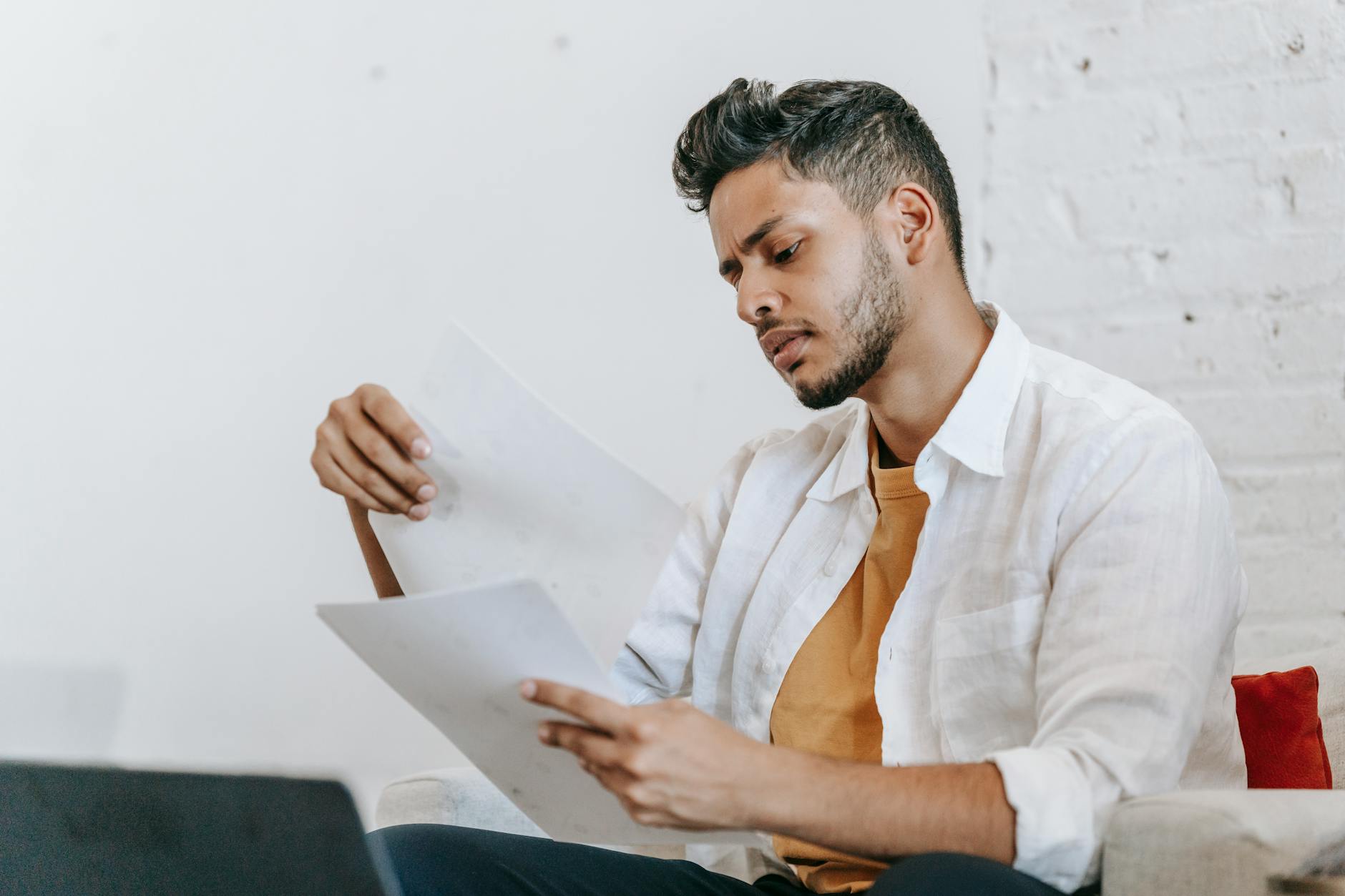 A man in a white shirt sitting on a couch, deeply focused on reading documents.