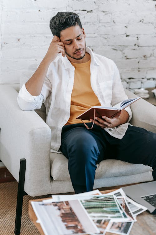 Free Concentrated young ethnic male wearing casual clothes reading notes in planner and touching face while sitting on cozy armchair Stock Photo