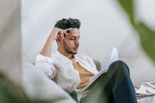 Concentrated ethnic male in casual clothes reading notes in diary while sitting on cozy sofa in living room