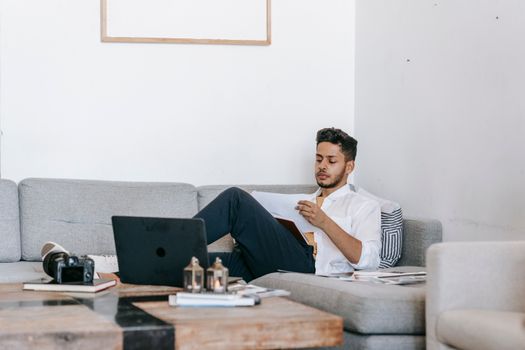 A young man with a beard using a laptop while sitting on a sofa at home, capturing a modern work-from-home lifestyle.
