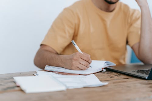 Free Crop anonymous male in casual wear working on netbook and taking notes in notepad while sitting at desk Stock Photo