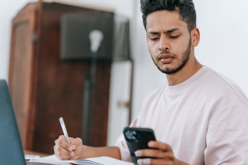 Concentrated ethnic male using mobile phone and taking notes in notebook while working on netbook at desk