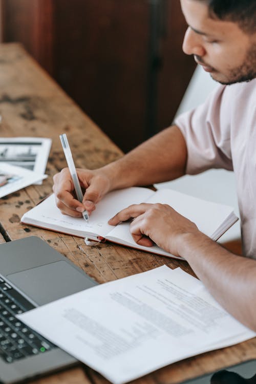 Free Crop focused man writing in copybook and using laptop Stock Photo