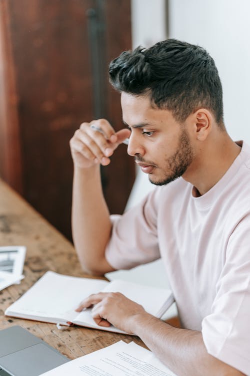 Concentrated ethnic male in casual wear working on netbook and sitting at desk with notebook