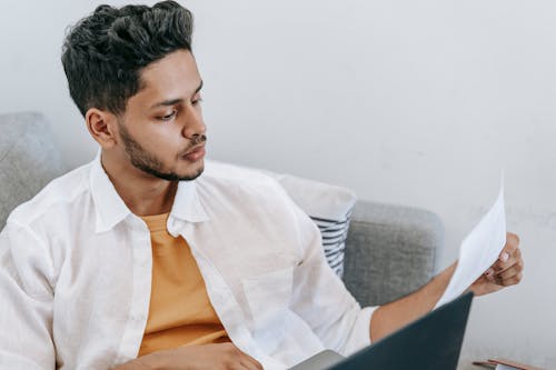 Serious ethnic man reading document and using laptop on sofa