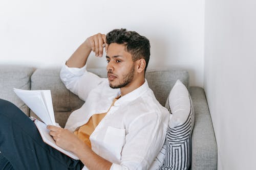 Concentrated young ethnic male wearing casual clothes reading documents and diary and touching head while resting on sofa and looking away