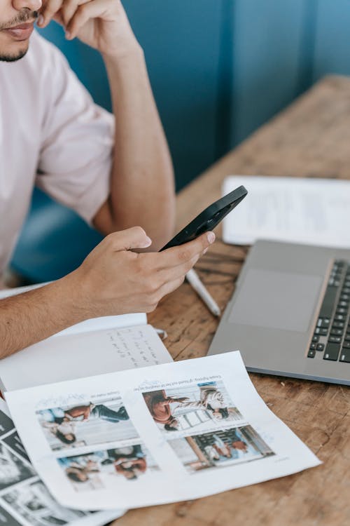 Crop focused male browsing mobile phone and working on netbook while sitting at desk with papers