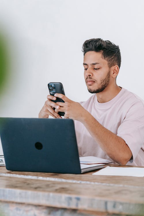 Free Thoughtful ethnic man browsing smartphone and working on laptop Stock Photo