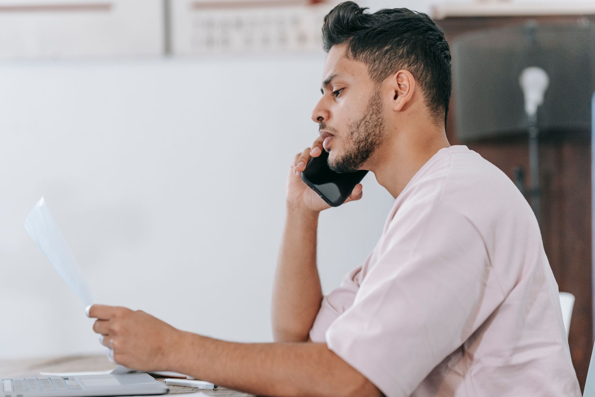 Side view thoughtful ethnic male in casual shirt reading document and talking on mobile phone while sitting at desk