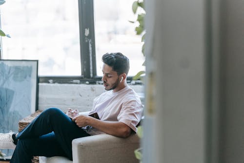 Pensive young ethnic male in casual wear writing in diary and listening to music via earbuds while sitting in cozy armchair