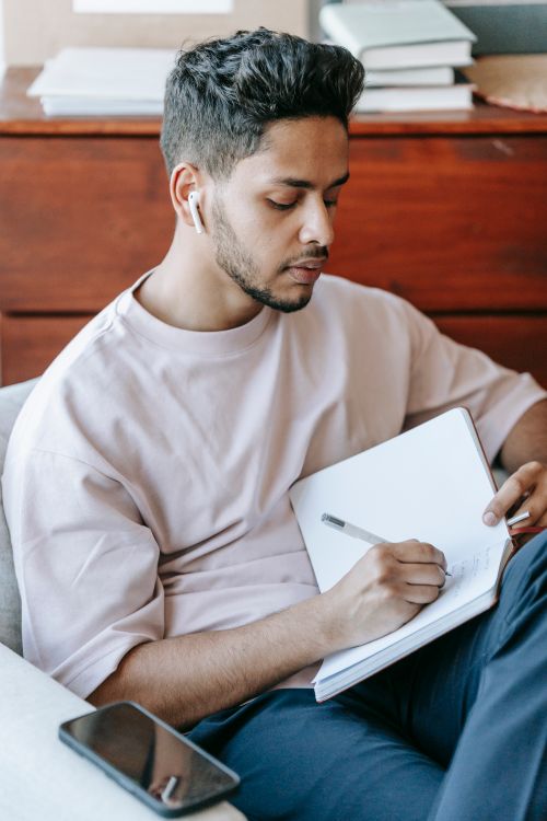 Free Pensive ethnic male taking notes in diary and listening to music via earbuds while sitting with legs crossed in soft armchair Stock Photo