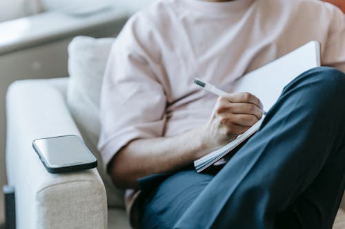 Free Crop unrecognizable male wearing casual outfit taking notes in planner and sitting with legs crossed in comfy armchair Stock Photo