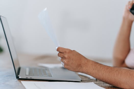 A man multitasking, talking on phone and reviewing papers while working on a laptop indoors.