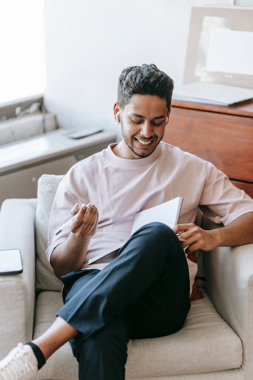 Happy young ethnic male wearing casual outfit taking notes in planner and listening to music in earbuds while sitting with legs crossed in cozy armchair