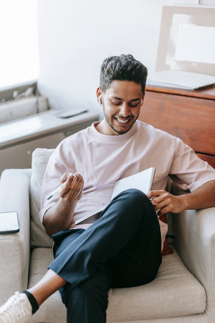 Cheerful Ethnic Man Writing In Diary And Listening To Music