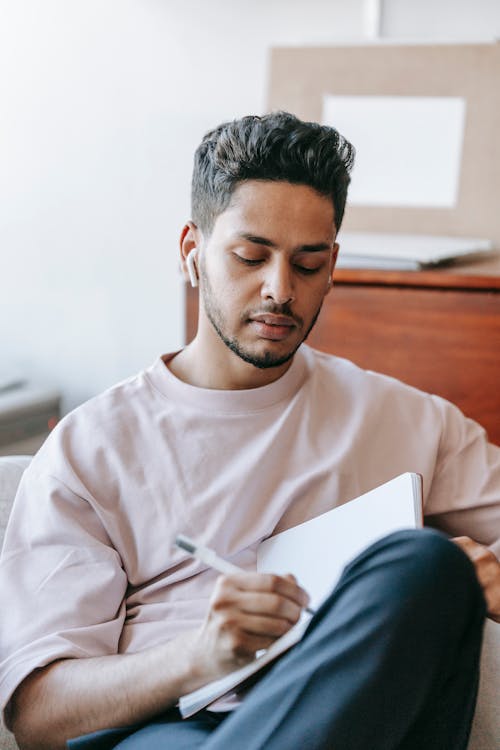 Pensive ethnic man in earbuds writing in notebook