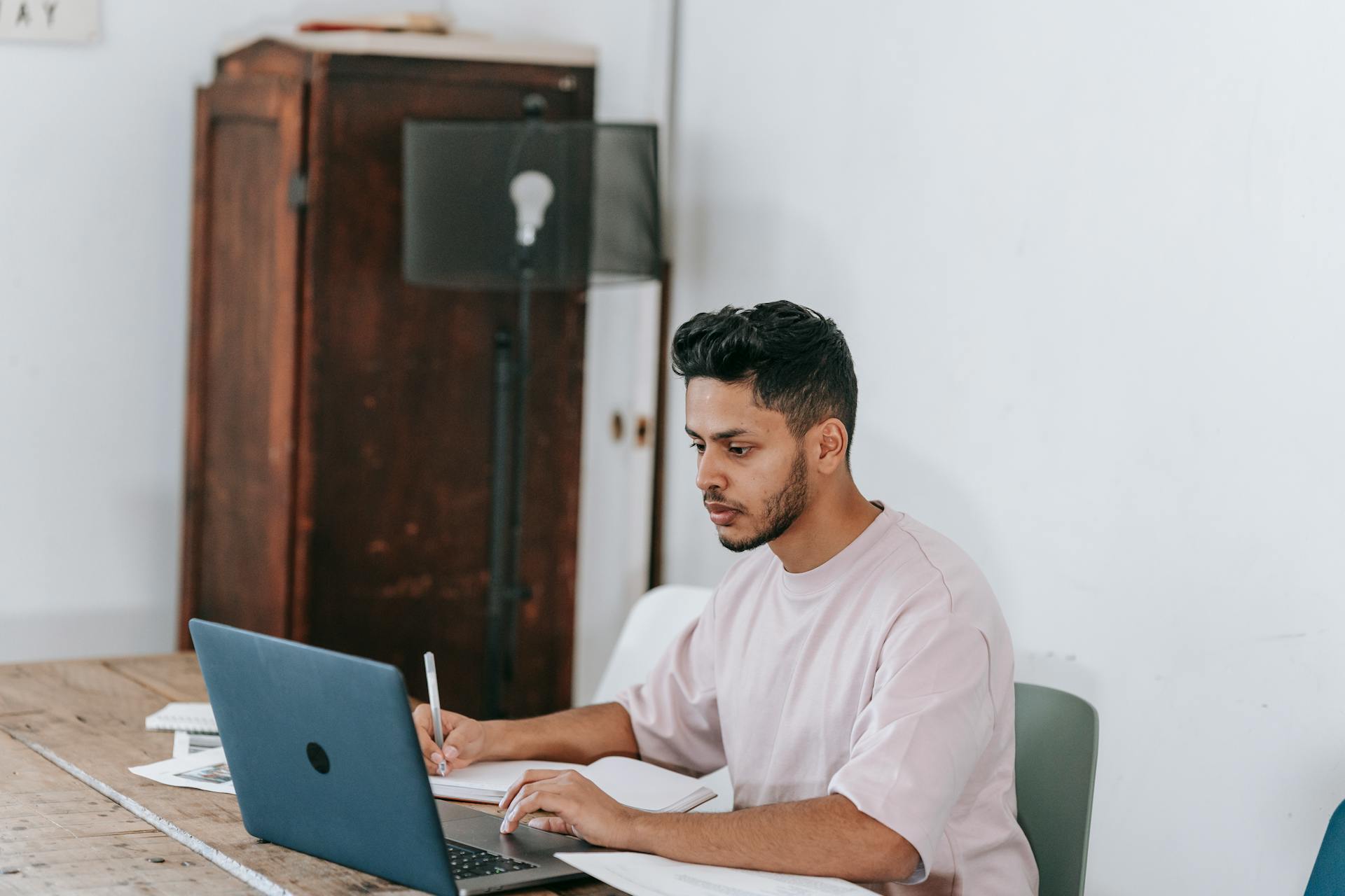 Focused young ethnic male in t shirt browsing netbook and taking notes in diary while working from home