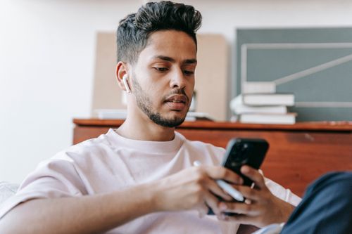 Free Emotionless young ethnic male in casual clothes and earbuds browsing mobile phone and sitting in light living room Stock Photo