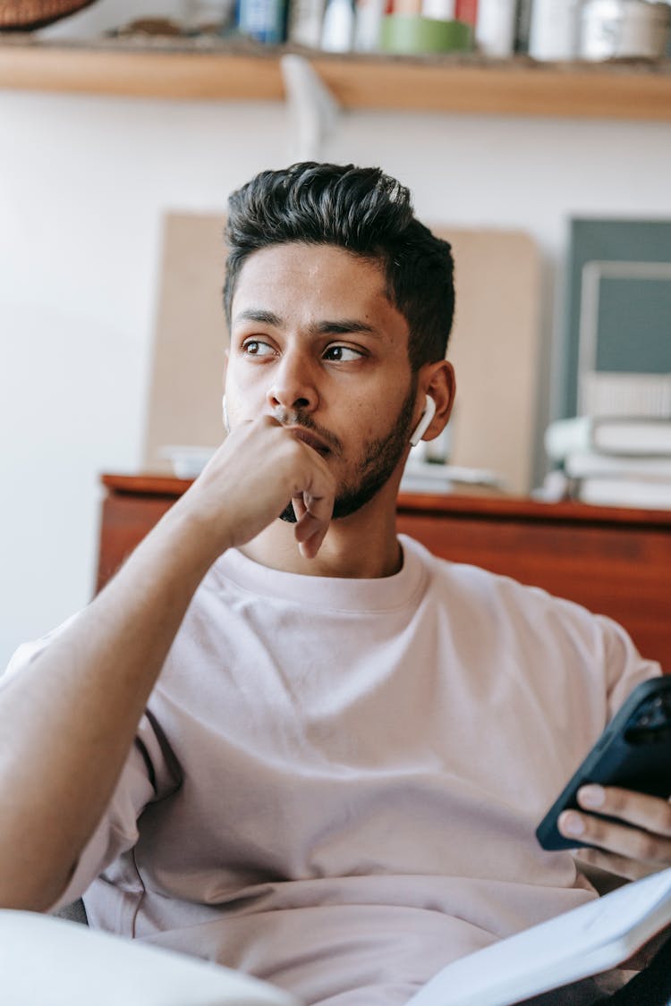 Thoughtful Ethnic Man In Earbuds Using Smartphone On Armchair