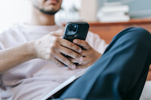 Free Crop bearded male in casual clothes browsing mobile phone and sitting in light living room Stock Photo