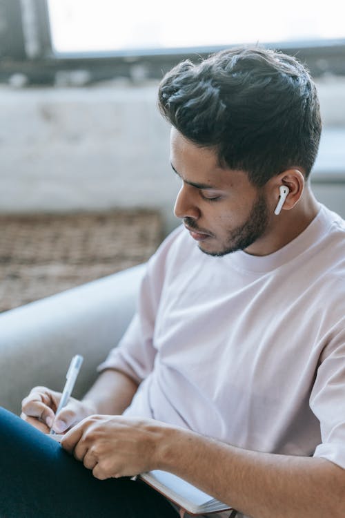 Concentrated young ethnic male wearing casual clothes taking notes in notepad and listening to audio via earbuds while sitting in armchair