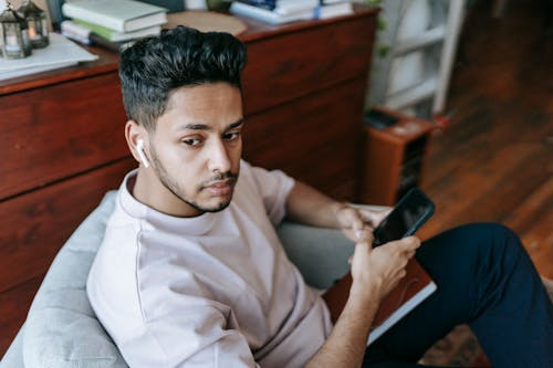 Young ethnic man in earbuds using smartphone in armchair
