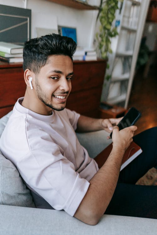 Free Side view happy young male using mobile phone and listening to music in earbuds while resting in cozy armchair Stock Photo