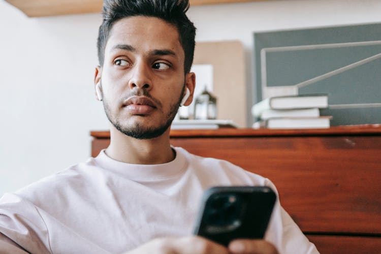 Pensive Indian Man Browsing Smartphone While Listening To Music