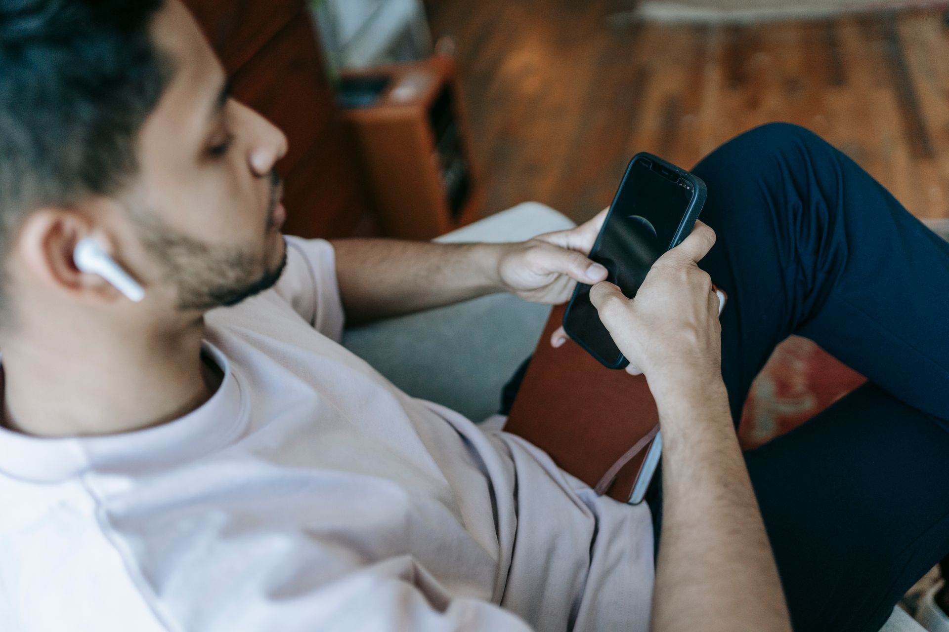 Young man using smartphone and wireless earbuds indoors, focusing on screen.