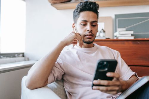 Free Serious Indian male listening to music in wireless earphones while browsing modern cellphone in armchair in light room at home Stock Photo