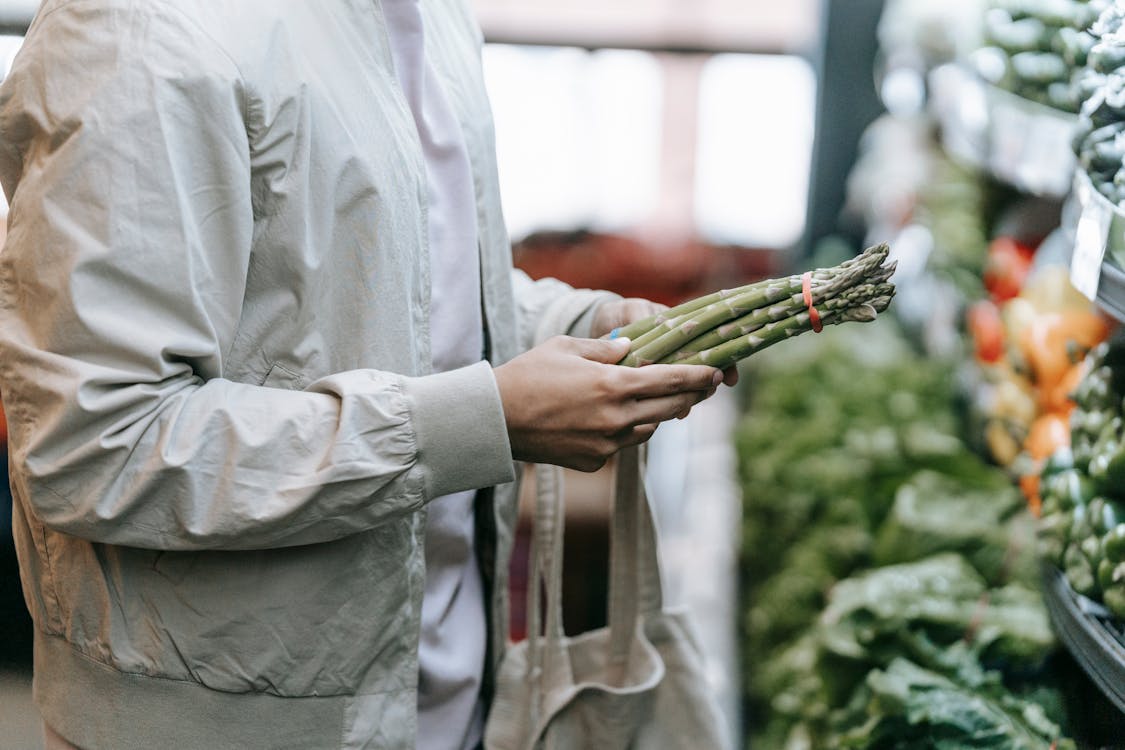 Free Crop faceless buyer with shopping bag picking green asparagus while standing near stall with fresh greens in supermarket on blurred background Stock Photo