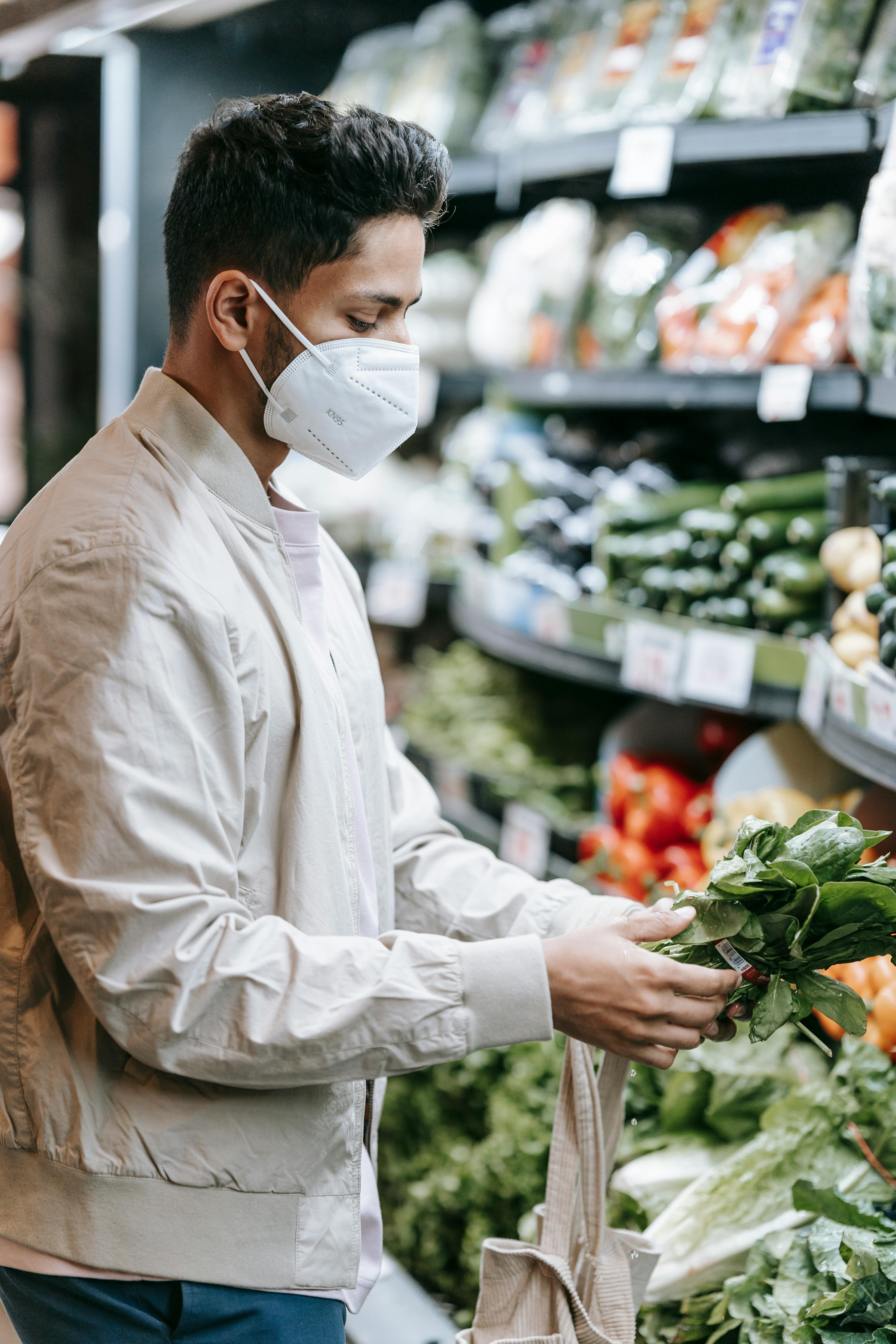 serious indian man in protective mask choosing greens in supermarket