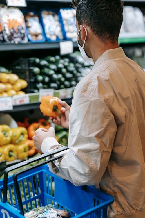 Free Back view of anonymous male buyer in protective mask picking ripe bell pepper while standing in vegetable department in supermarket Stock Photo