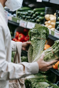 Crop anonymous buyer in protective mask with shopping bag choosing fresh greens while standing near stall with vegetables in grocery store