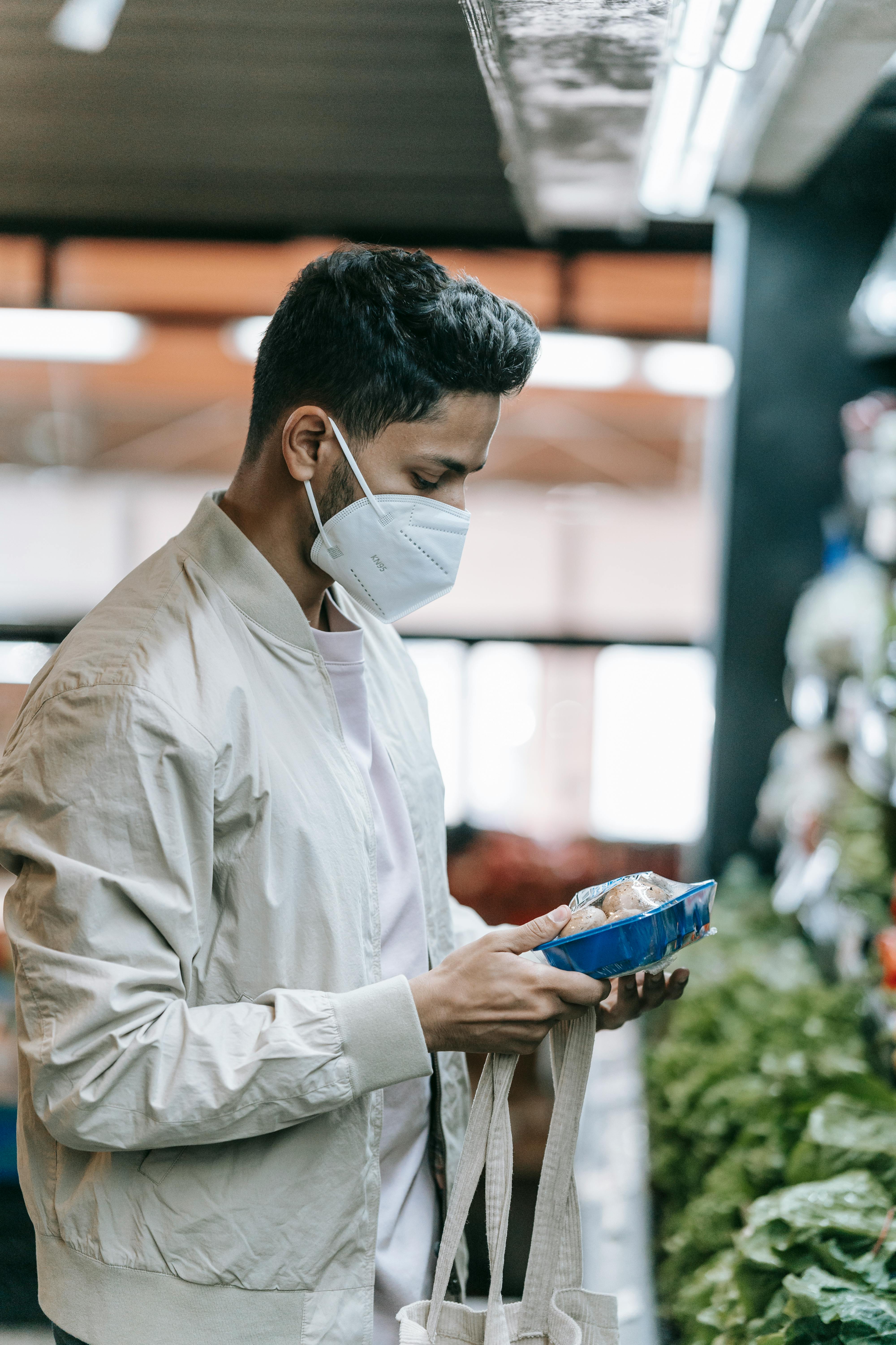 focused indian man in medical mask with mushrooms in supermarket