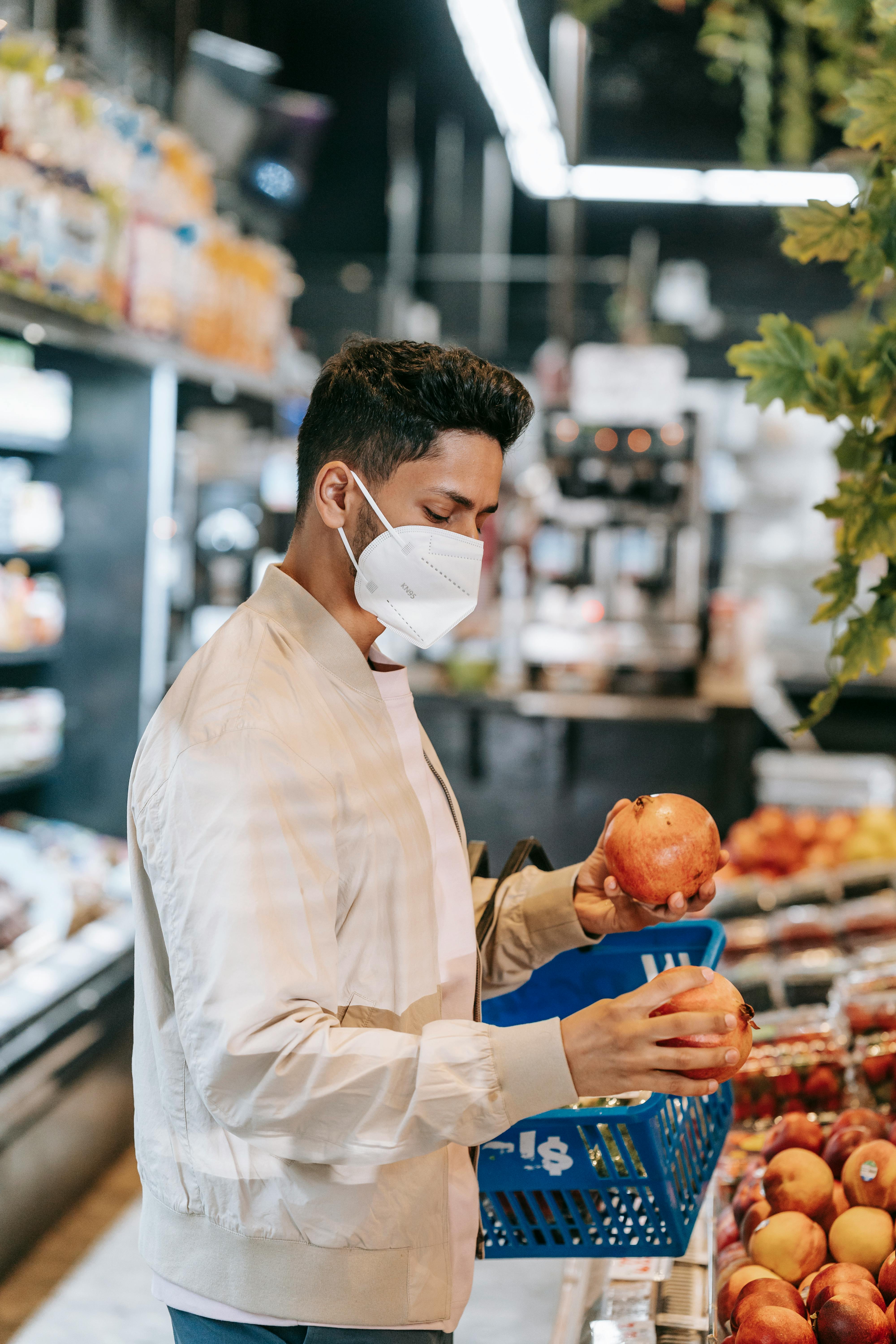 Side view of anonymous young ethnic male buyer with dark hair in stylish jacket and medical mask choosing fresh pomegranates while standing near tables with assorted fruits in modern supermarket