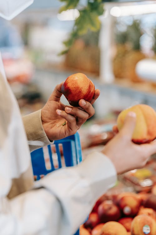 Side view of crop faceless male customer in casual clothes and medical mask choosing between nectarine and peach in modern grocery market