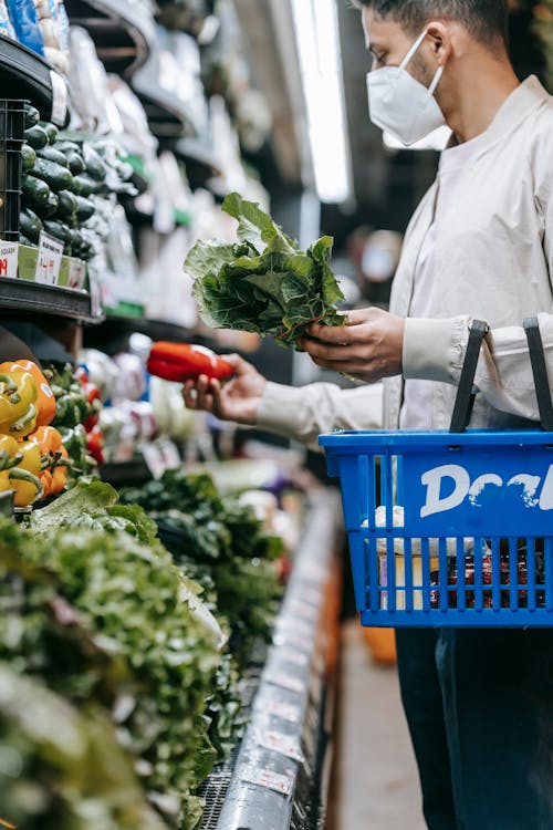 Side view of crop young ethnic male buyer in casual clothes and protective mask holding basket and choosing fresh spinach and vegetables during shopping in supermarket