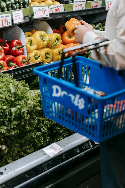 Free Side view of crop faceless buyer in casual clothes carrying shopping basket while choosing assorted colorful bell peppers in supermarket Stock Photo
