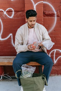 Trendy ethnic male sitting on bench with fresh vegetables between eco bag and brick wall on street