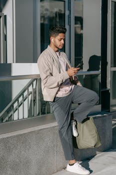 Stylish ethnic male with natural bag listening to music from earbud while browsing internet on cellphone in city