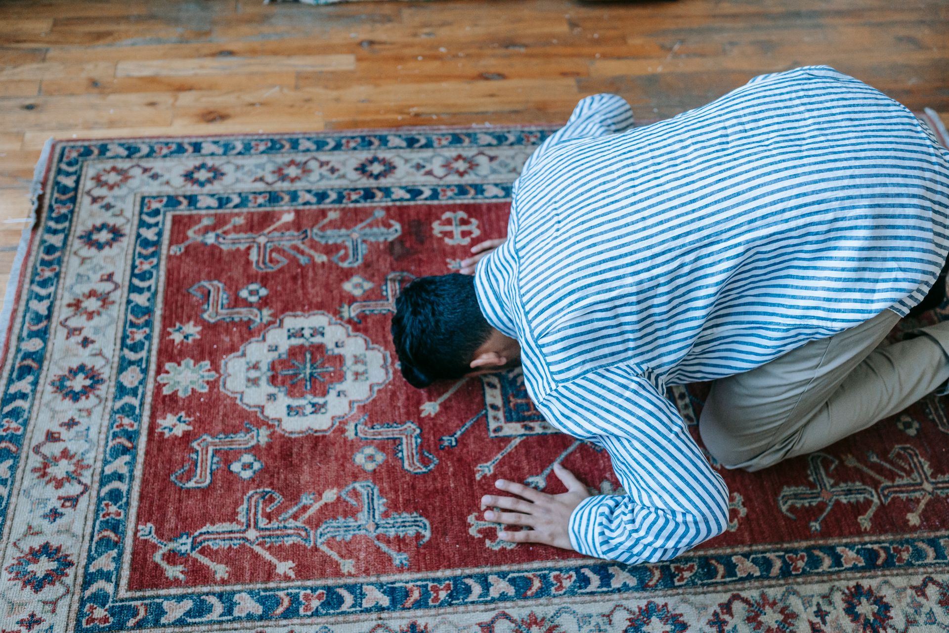 A Muslim man in striped clothing prays on a traditional decorative rug indoors.